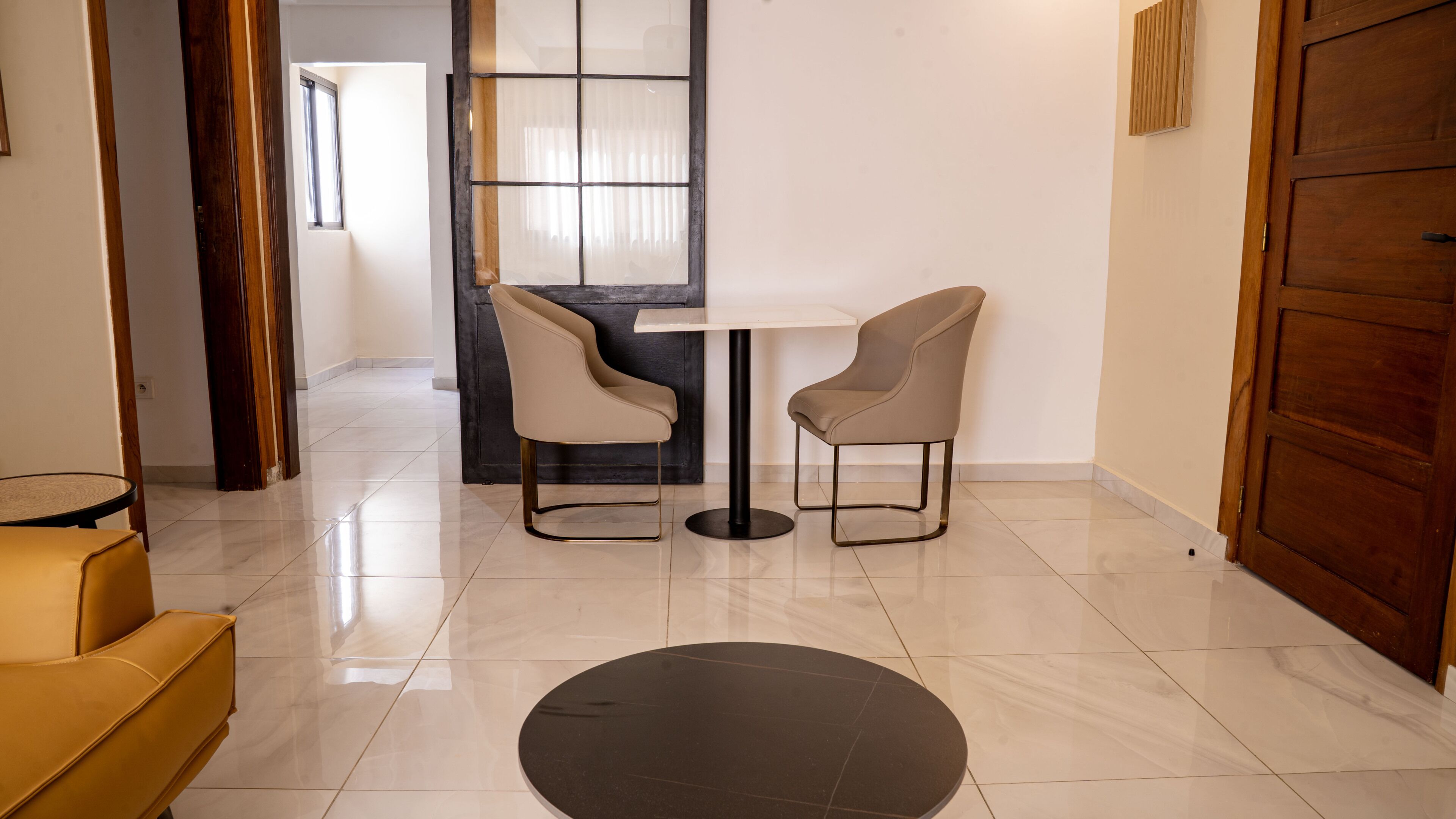 A modern interior with a round black rug, two grey chairs, a small round table, and a large window with blinds.