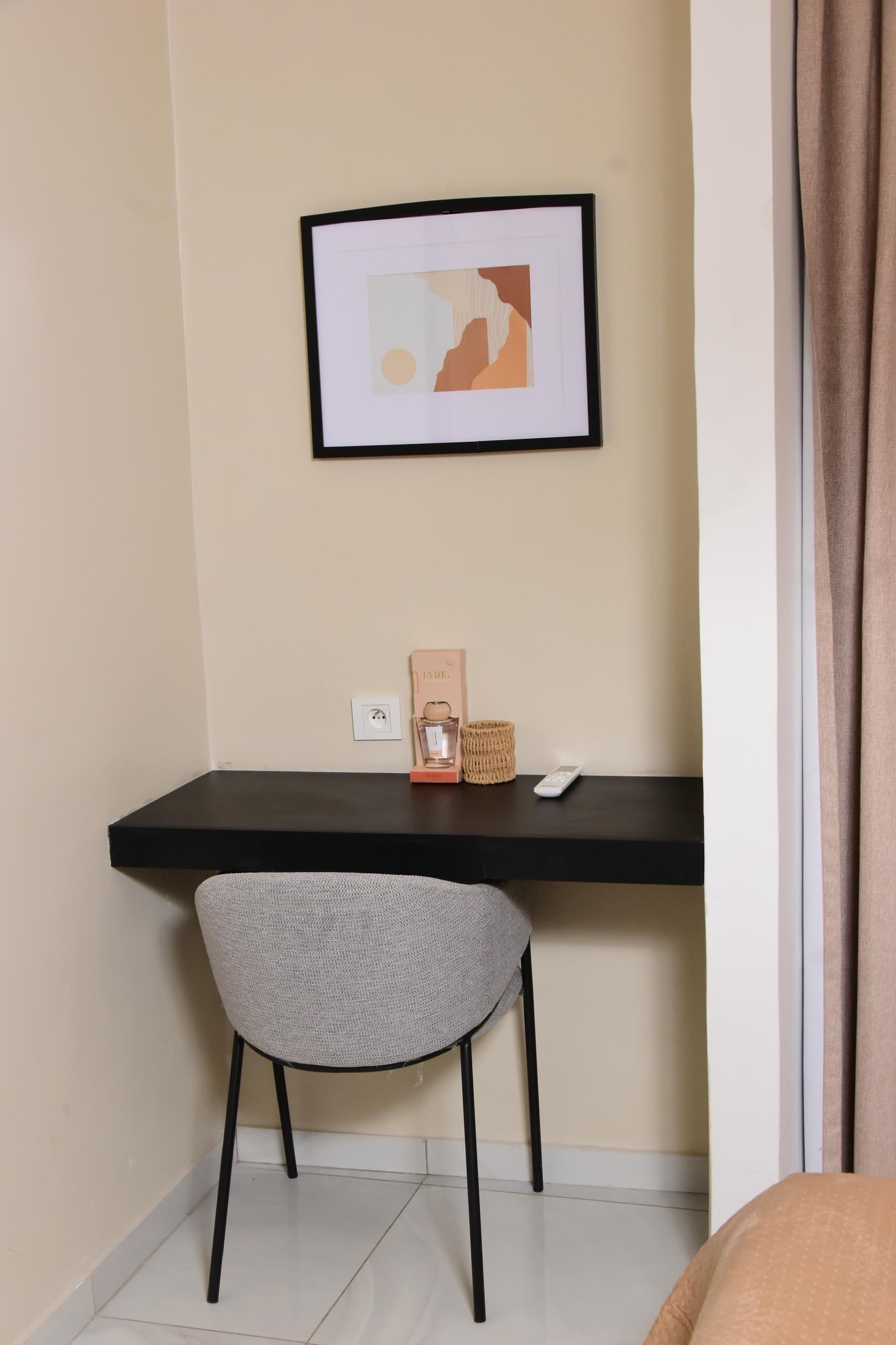 Ein kleines Zimmer mit einem Schreibtisch, einem Stuhl, einem gerahmten Bild, einer Fernbedienung und einer Dekoration auf dem Schreibtisch.