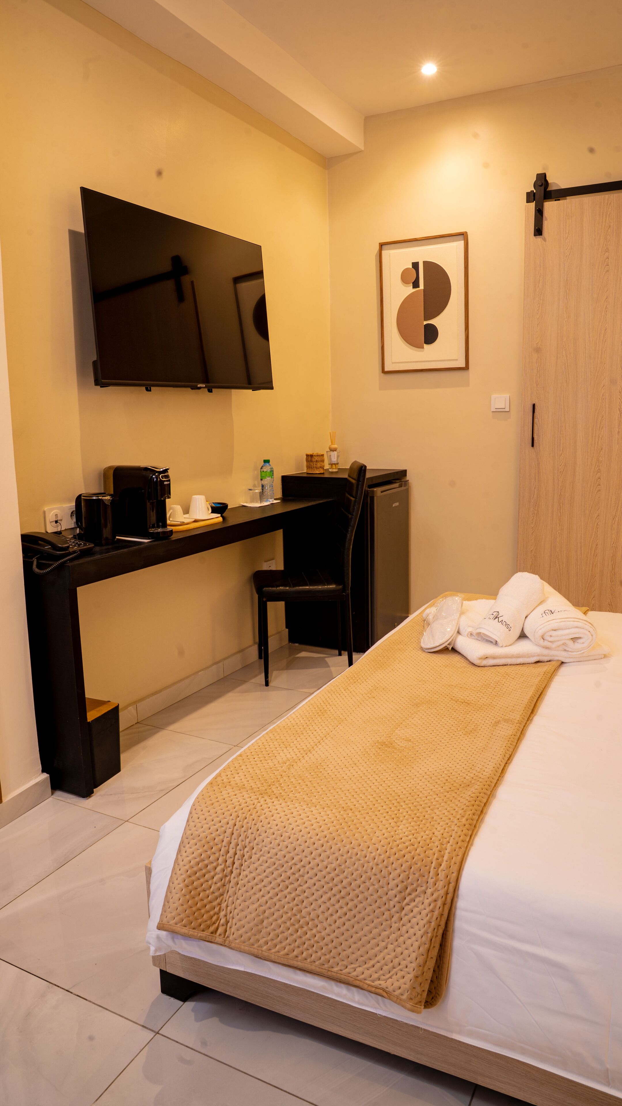 Habitación de hotel con cama, escritorio con silla, televisor, un cuadro enmarcado y armario.