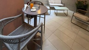 A patio area with a round table, two wicker chairs, a white sofa, a potted plant, and a textured wall. - Kadiss Hotel (Dakar)