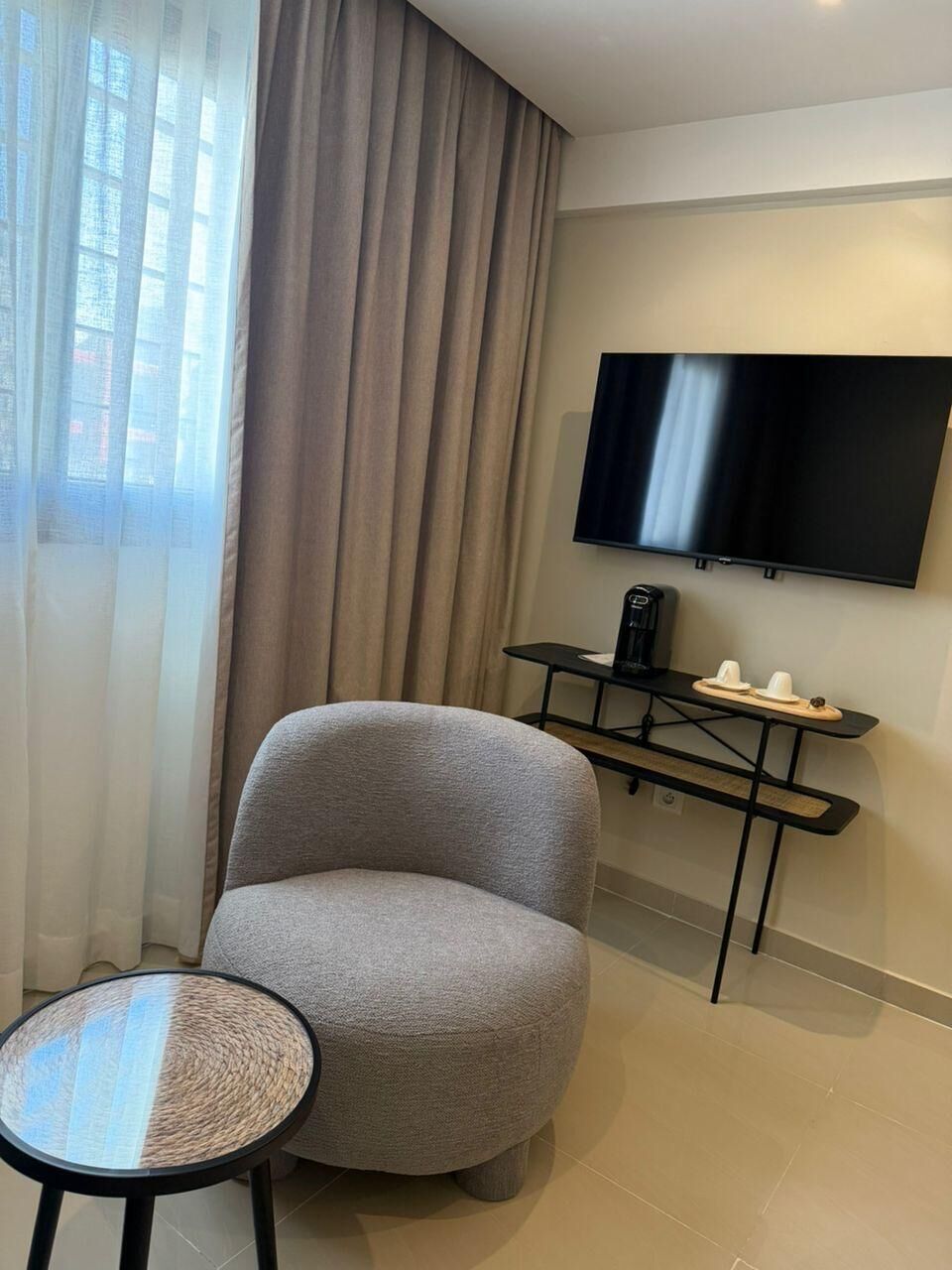 A modern hotel room with a grey armchair, a small round table, a flat-screen TV mounted on the wall, and a coffee machine on a black table.