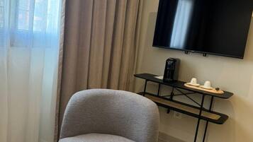 A modern hotel room with a grey armchair, a small round table, a flat-screen TV mounted on the wall, and a coffee machine on a black table.