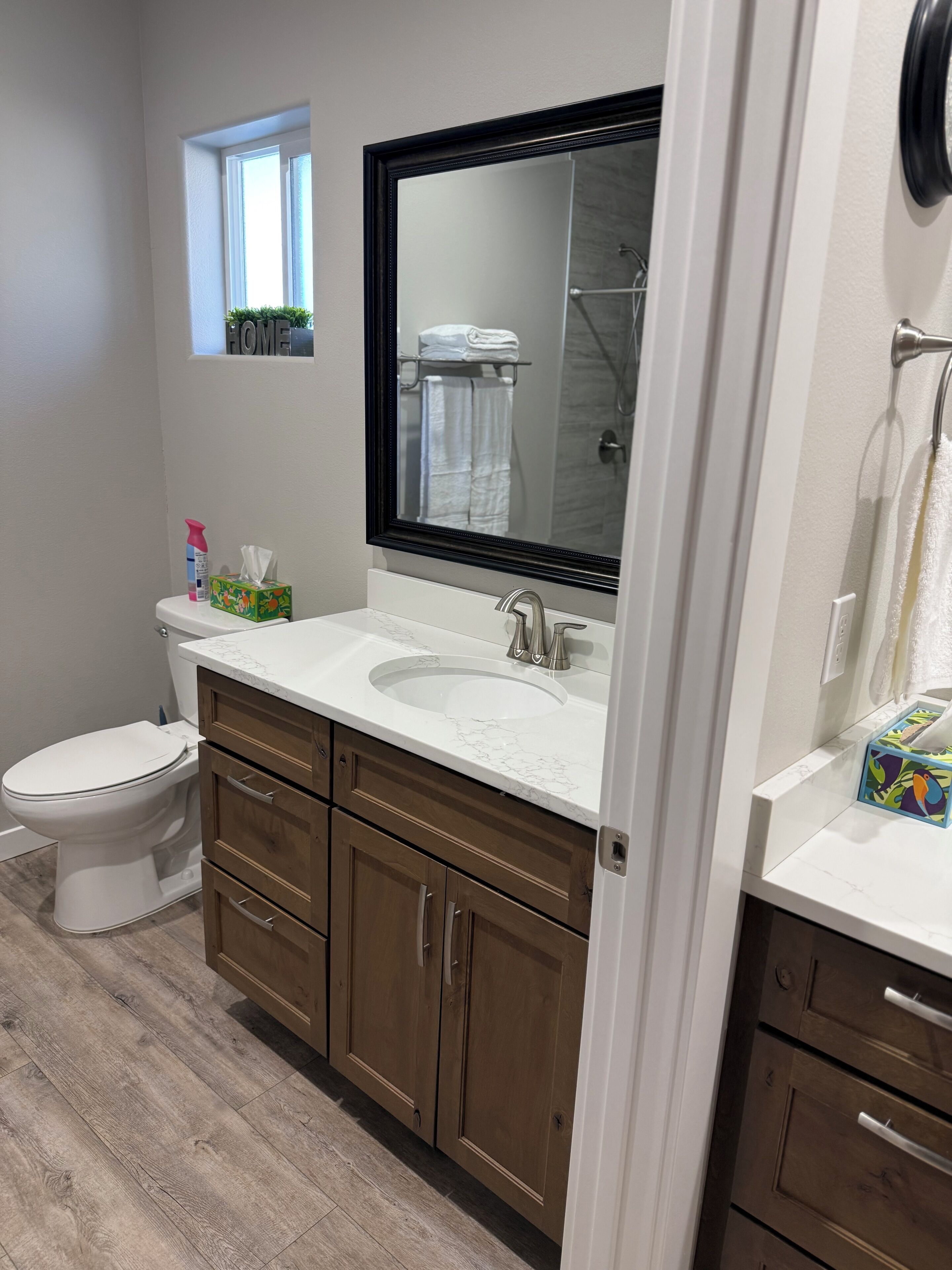 A bathroom with a toilet, a sink with a large mirror, wooden cabinets, and a window with a view of the outside.