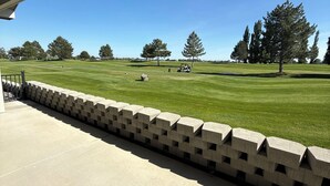 Blick von einer überdachten Veranda auf einen Golfplatz bei blauem Himmel.