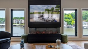 Een moderne woonkamer met een open haard, een flatscreen televisie en grote ramen met uitzicht op een waterpartij en gebouwen.
