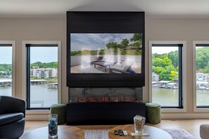 Una sala de estar moderna con chimenea, televisor de pantalla plana y ventanas amplias con vistas a un cuerpo de agua y edificios.