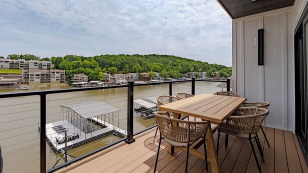 Een balkon met een tafel en stoelen, met uitzicht op een rivier, met gebouwen en groen op de achtergrond.
