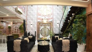 A luxurious lobby with a central decorative wall, two black sofas with gold legs, a round coffee table, and a staircase with greenery.