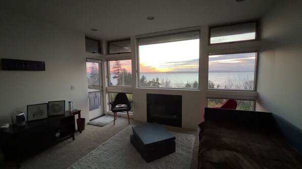 A living room with a fireplace, large windows with a view of the water, a sofa, and a coffee table. - Modern private ocean-view 2-bedroom mother-in-law with hot tub, walk to beaches. (Seattle)