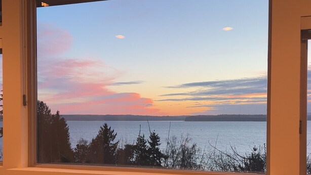 Huoneessa on takka, suuri ikkuna, josta avautuu näkymä vesistöön ja auringonlaskuun, sekä matolla peitetty lattia.