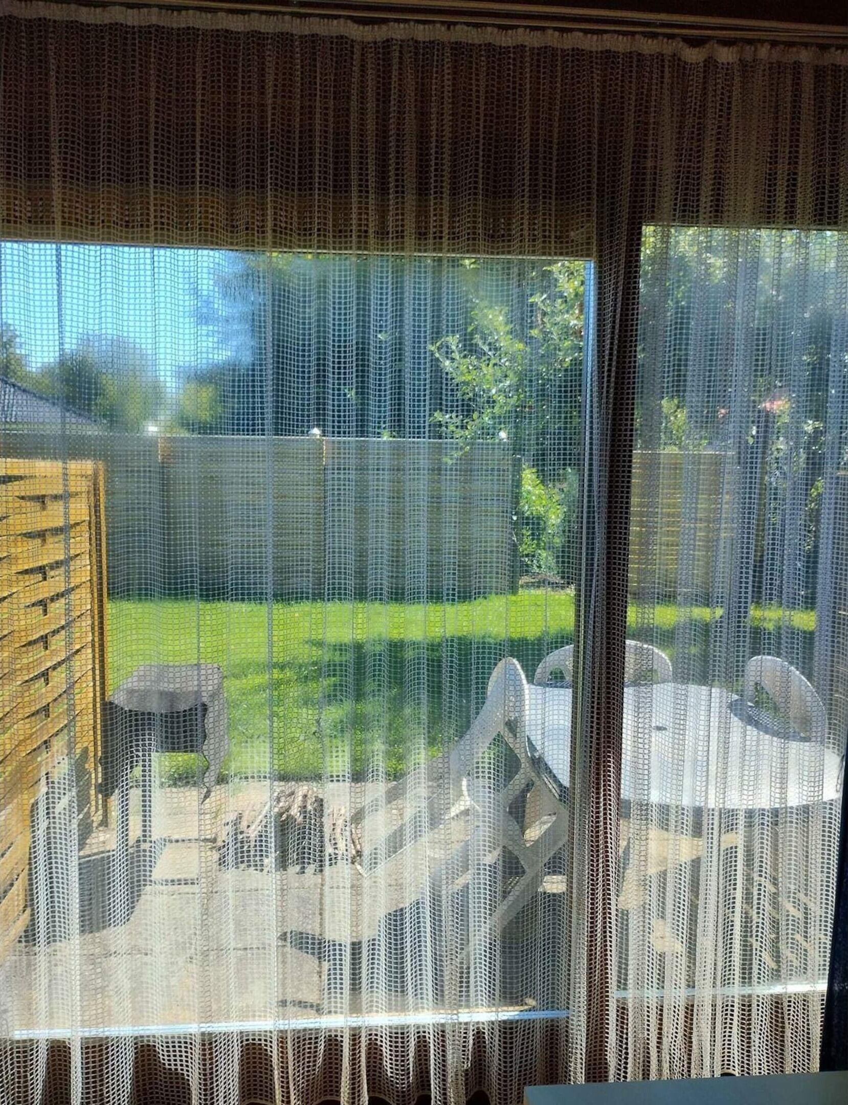 A view through a window with a mesh screen, a wooden fence, and a grassy yard with a table and chairs.
