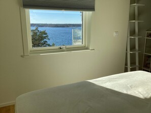 Quarto com janela dando para a água, cama branca e estante branca.