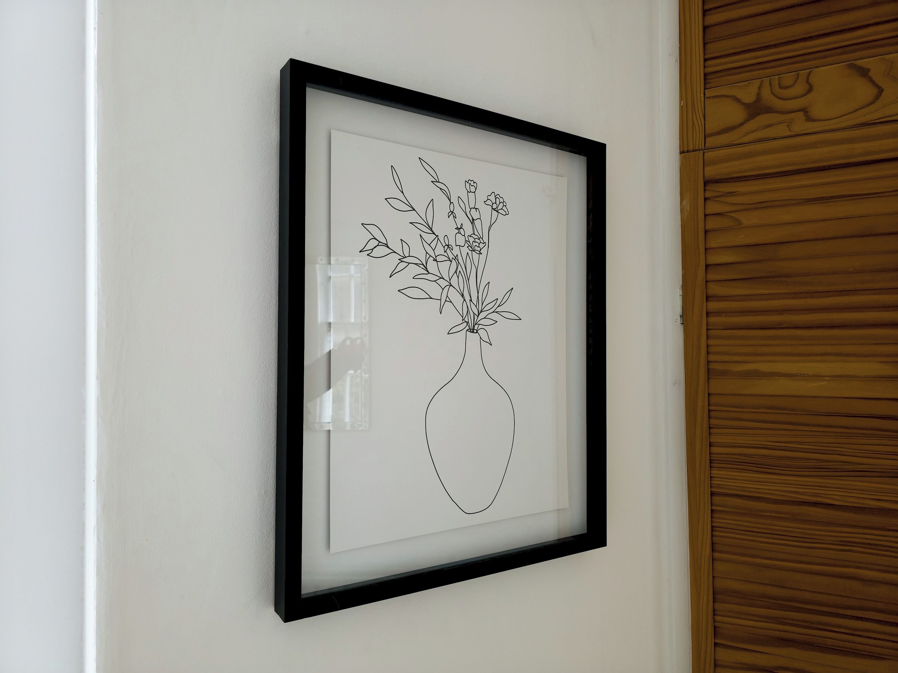 A framed black and white line drawing of a vase with flowers on a white wall.