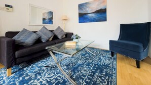 A living room with a black sofa, a glass coffee table, a blue patterned rug, a blue armchair, and two paintings on the wall. - Stylish Flat Near Whitechapel - Pass the Keys (London)