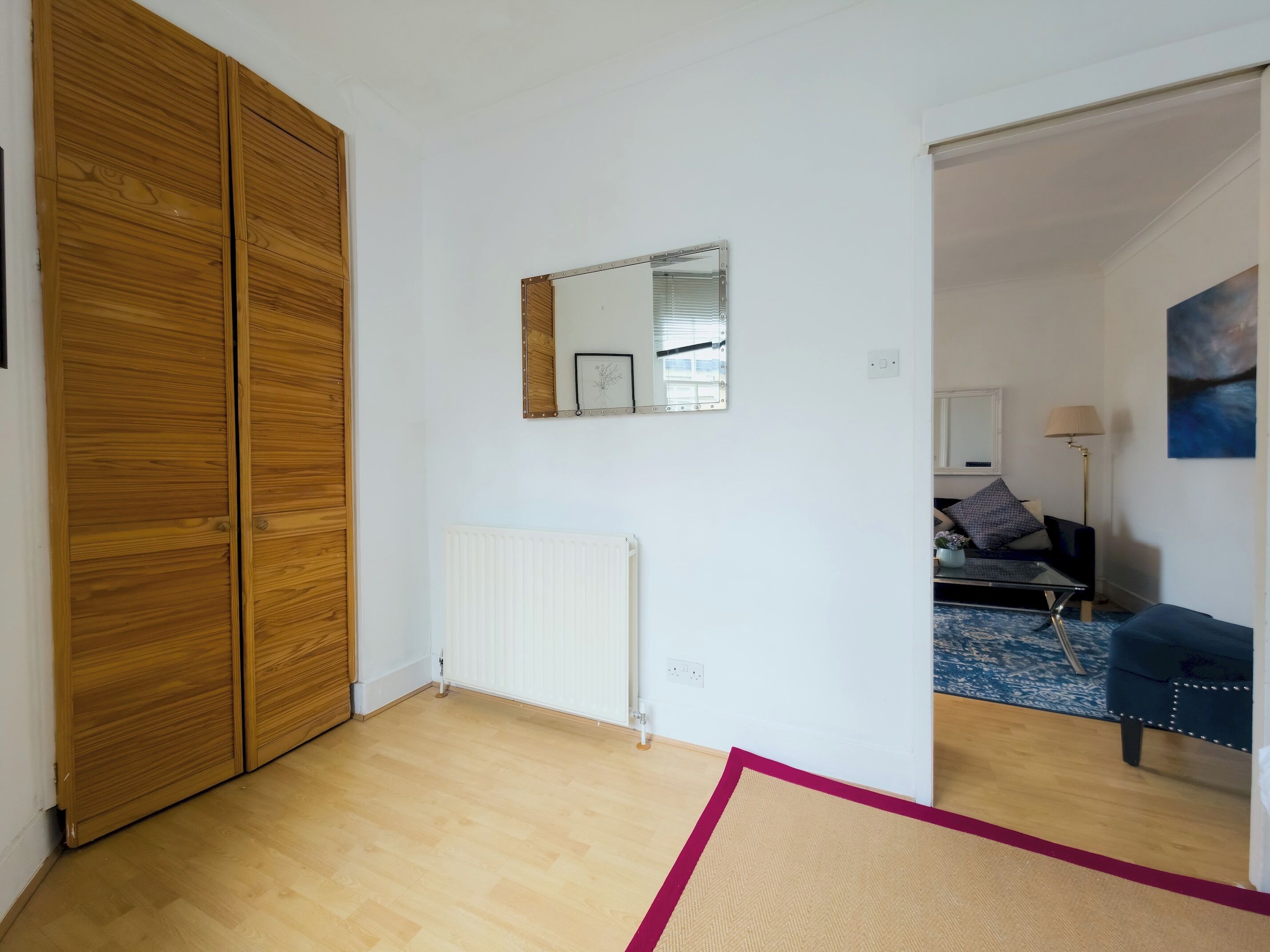 A room with a wooden wardrobe, a radiator, a mirror, a bed with a blue headboard, and a lamp.