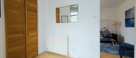 A room with a wooden wardrobe, a radiator, a mirror, a bed with a blue headboard, and a lamp.