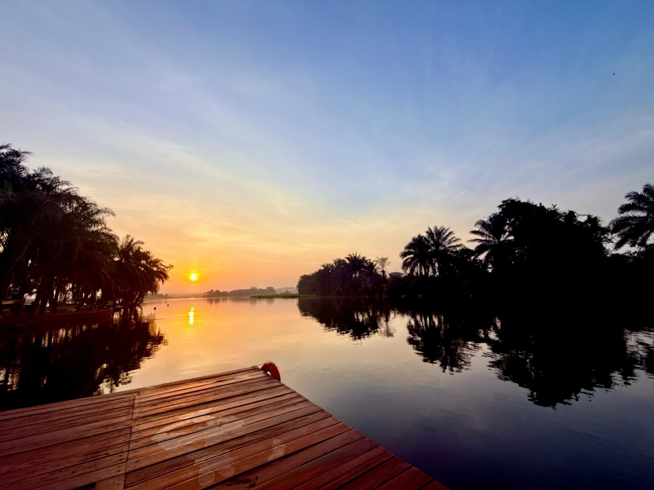 A wooden dock extending into calm water with palm trees on the banks, reflecting the sunset.
