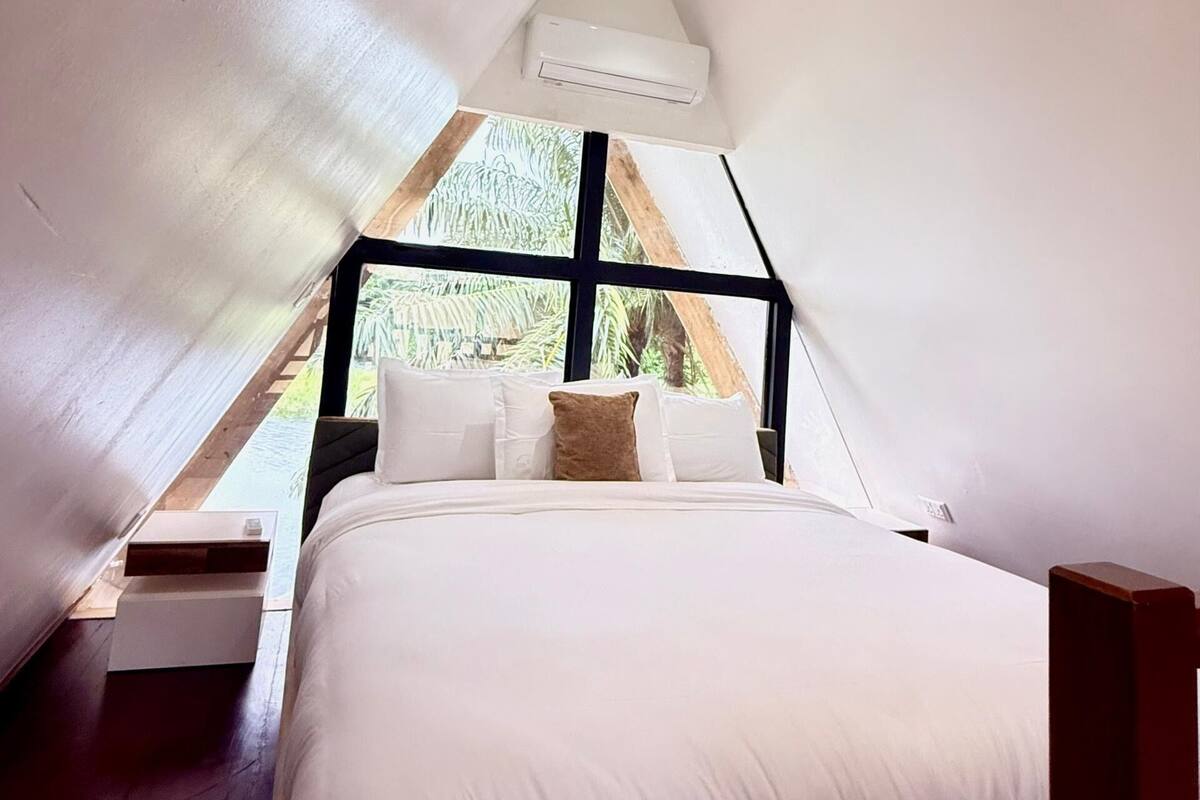 Un dormitorio con cama, almohadas, una mesita de noche, una ventana y un equipo de aire acondicionado.