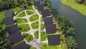 An aerial view of a resort with multiple dark-roofed cabins arranged in a semi-circular pattern around a central paved area, surrounded by lush greenery and a body of water. - Palm Island Resort (Akosombo)
