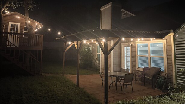 Uma área de varanda coberta com luzes pendentes, um deck de madeira e uma mesa de jantar com cadeiras.