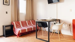 Una habitación con sofá, una mesa y sillas, televisor montado en la pared y ventana con cortinas.