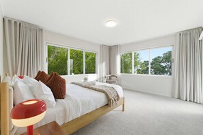 A modern bedroom with a large bed, a bedside table, a chair, and a window with a view of trees. - Spacious 4BR Birkenhead Retreat | 3 Living Areas (Auckland North Shore)