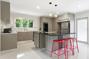 A modern kitchen with a central island, red bar stools, and a view of greenery outside. - Spacious 4BR Birkenhead Retreat | 3 Living Areas (Auckland North Shore)