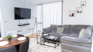 Een moderne woonkamer met een grijze bank, een salontafel, een aan de muur bevestigde televisie en een eethoek met een tafel en stoelen.