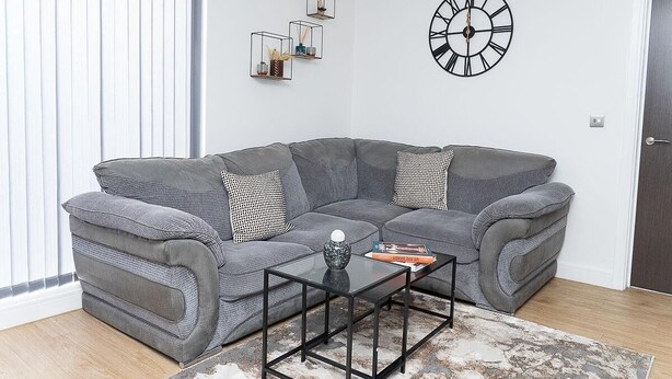 Een moderne woonkamer met een grijze modulaire bank, een glazen salontafel, een zwarte klok en een geometrisch wanddecoratie.