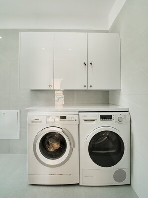A laundry area with two front-loading washing machines, a white cabinet above, and a tiled wall. - Central sunset view #2 (Himarë)