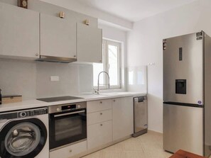 A compact kitchen with a washing machine, oven, dishwasher, and a stainless steel refrigerator. - Central sunset view #1 (Himarë)
