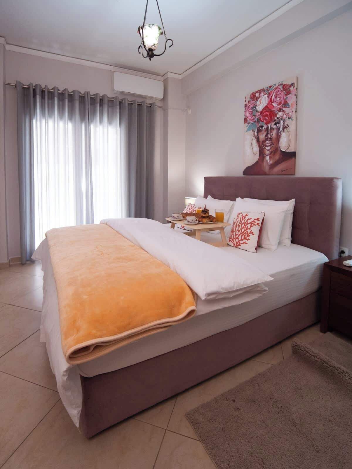 Una camera d'albergo con un letto, un comodino, un tavolino di fianco al letto, un quadro appeso al muro e una finestra con tende.