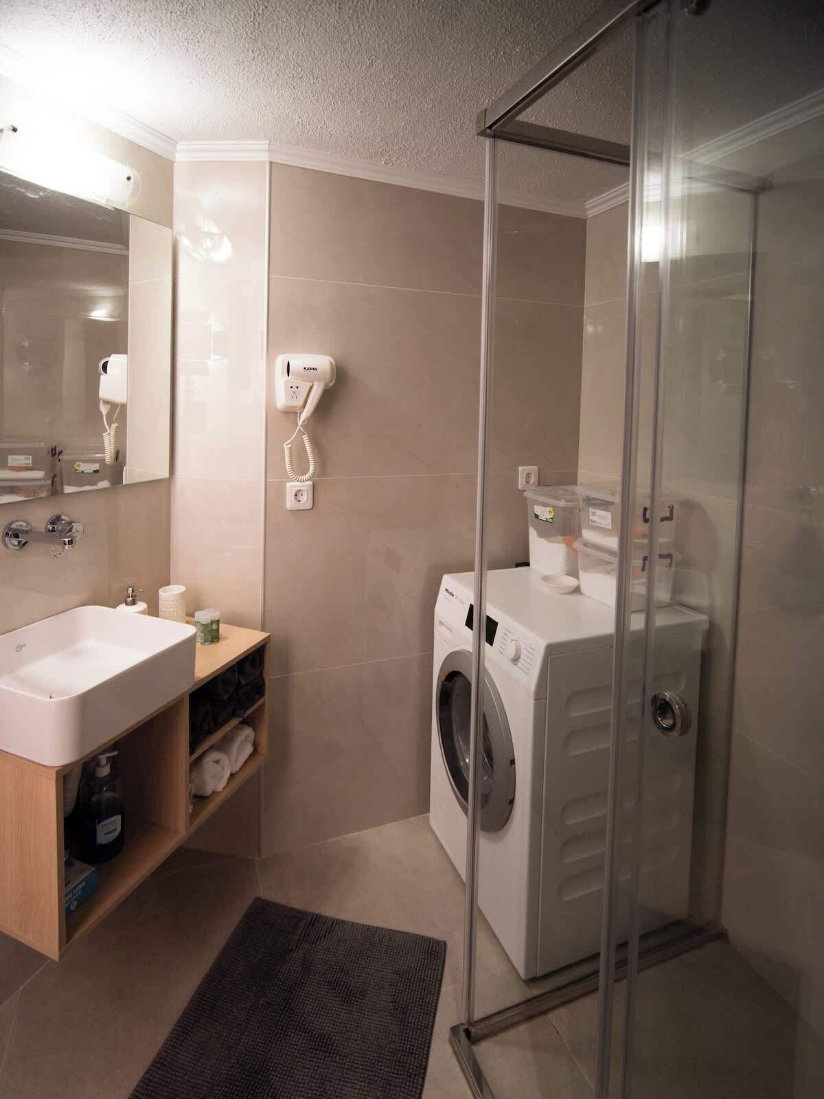 Une salle de bain moderne équipée d’un lavabo, d’un miroir, d’un sèche-cheveux et d’une douche séparée par des parois en verre.