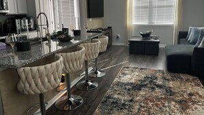 Eine moderne Küche mit Barbereich, einem Essbereich mit Barhocker, einer Couch und einem großen Fenster mit Jalousien.