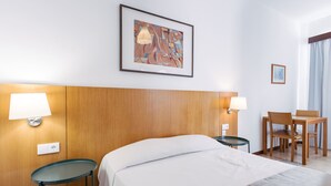 A hotel room with a bed, two bedside lamps, a desk with a chair, and a framed abstract painting on the wall. - Economy Studio Apartment in the heart of Funchal. 2' walk from Cathedral. (Funchal)