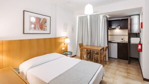 A hotel room with a bed, a desk, a chair, a small table, a lamp, a framed picture, a window with curtains, a kitchenette, and a fire extinguisher. - Economy Studio Apartment in the heart of Funchal. 2' walk from Cathedral. (Funchal)