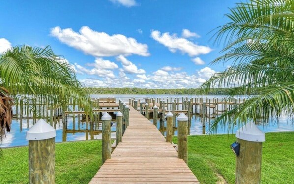 A wooden pier leading to a dock with a clear view of a lake and palm trees.