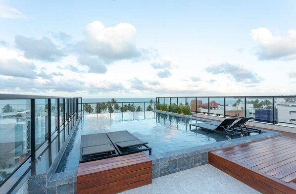 A rooftop infinity pool with lounge chairs, wooden deck, and a view of the city skyline.