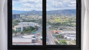 Un ventanal grande con cortinas blancas, con vistas a un paisaje urbano que incluye edificios, calles y montañas a lo lejos.