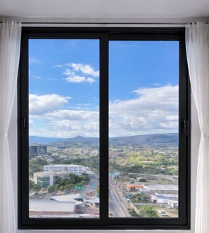 Ein großes Fenster mit weißen Vorhängen, das einen Blick auf eine Stadtlandschaft mit Gebäuden, Straßen und fernen Bergen bietet.