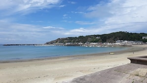 Une plage côtière, avec un ciel dégagé, une mer calme et une colline rocheuse en arrière-plan, sur laquelle se dressent des bâtiments.