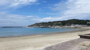A coastal beach with a clear sky, calm sea, and a rocky hill with buildings in the background.
