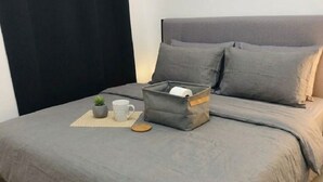 Una cama bien hecha con ropa de cama gris, un cesto de almacenamiento gris, una pequeña planta en maceta y una taza blanca sobre un tapete tejido.