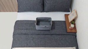 Una cama con ropa de cama gris, una almohada gris, un bolso gris, una bandeja de madera con una planta y un cuadro enmarcado.