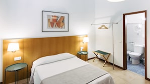 Ett hotellrum med en säng, sänglampor, ett inramat konstverk, ett litet bord, en hopfällbar stol och ett badrum som syns genom en öppen dörr.