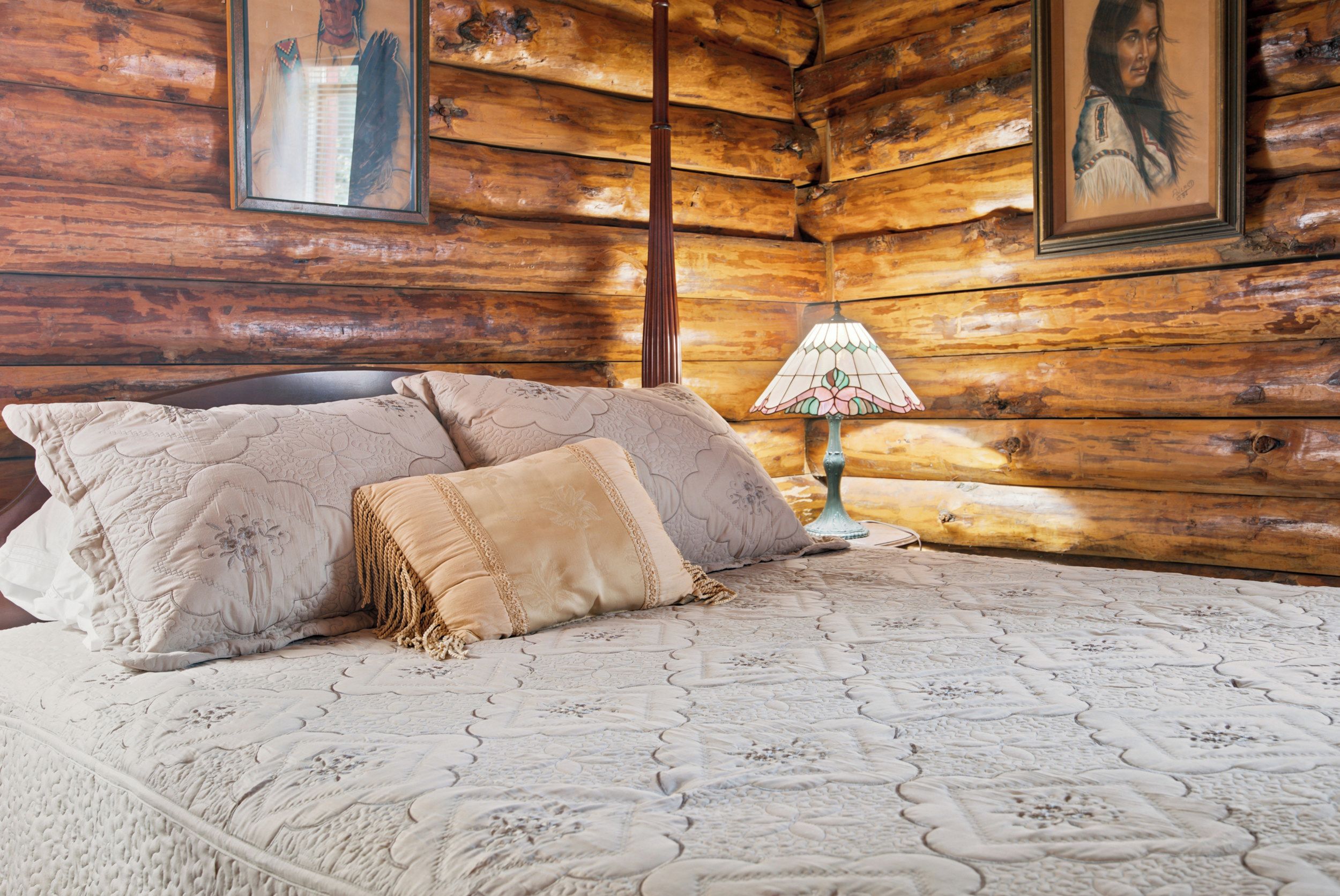 A wooden cabin interior with a bed, pillows, a lamp, and framed artwork on the walls.