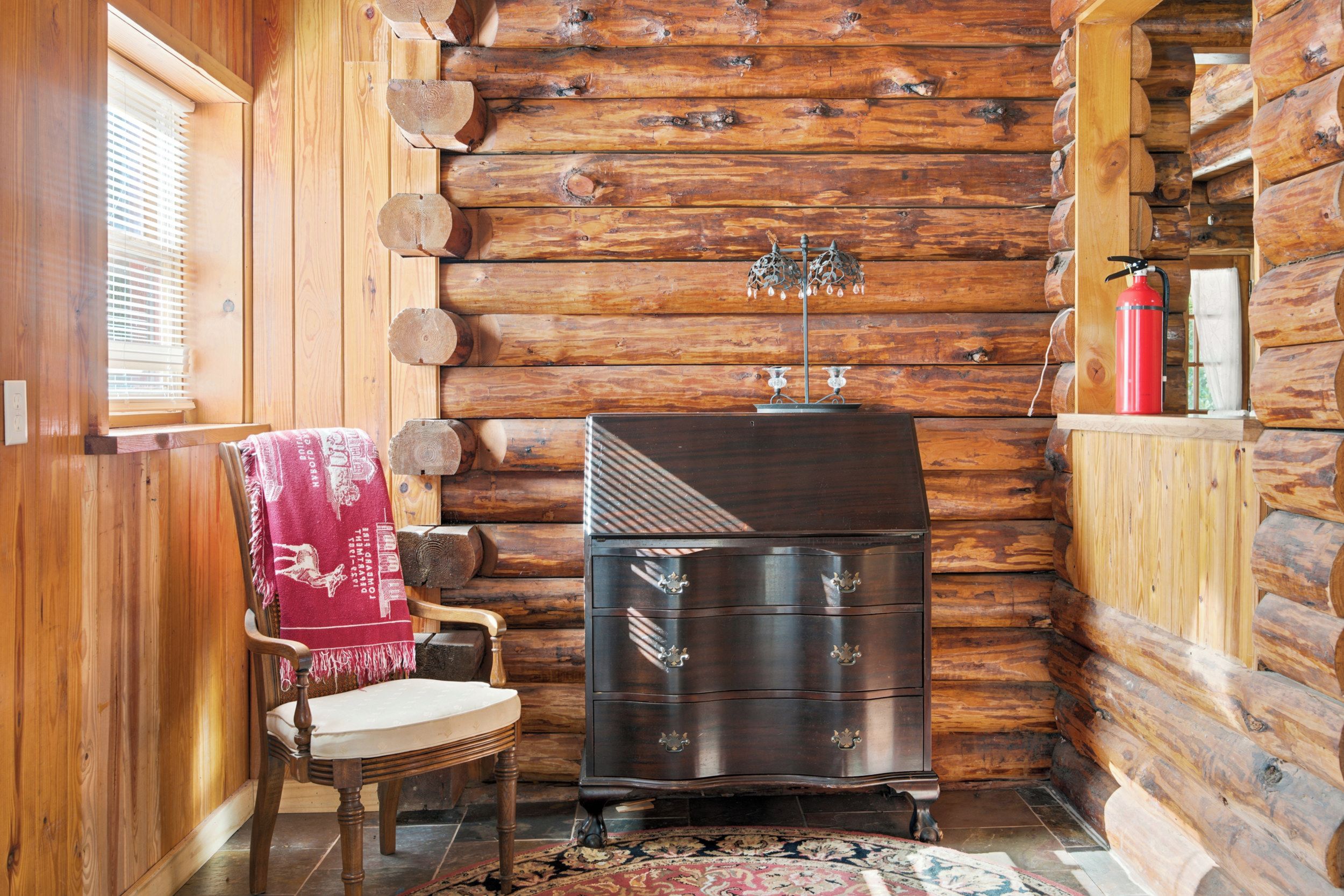 A log cabin interior with a wooden chair, a dark wooden dresser, and a window with blinds.