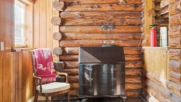 A log cabin interior with a wooden chair, a dark wooden dresser, and a window with blinds.