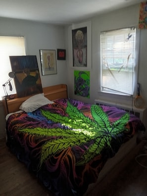 A bedroom with a bed, a desk, a chair, a window with blinds, and artwork on the walls.