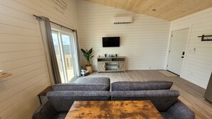 A modern living room with a wooden ceiling, a grey sofa set, a wooden coffee table, a TV mounted on the wall, and a sliding glass door leading to an outdoor area.